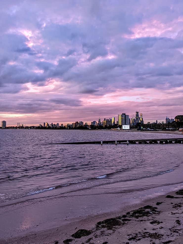 The sky lights up in shades of purple and pink behind moody looking clouds as you look across the coastline of St Kilda to the city lights and skyscrapers of Melbourne CBD