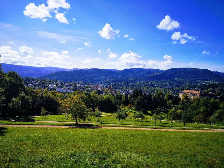 Walking paths cross lush green parkland, dotted with trees. The town of Baden Baden can be seen in the distance, surrounded by mountains of forest.