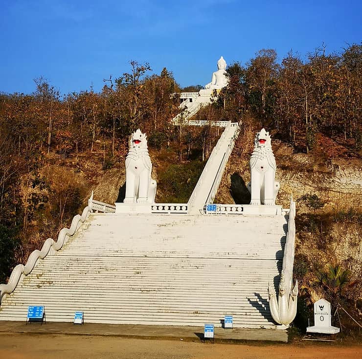 A grand white staircase, with two white lion statues on either side leads up through the trees to a big white Buddha on the top of a hill in Pai, Thailand