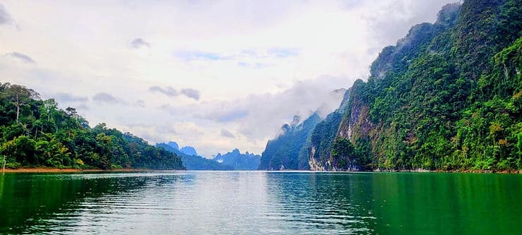 The vast Cheow Lan lake in Khao Sok National park. On the right, huge limestone cliffs rise from the water. On the left side, the jungle sits behind a thin sandy shoreline. 