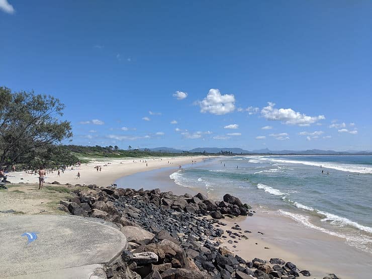 The white sandy Belongil beach in Byron Bay spans into the distance, backed by trees as waves crash gently onto the shore