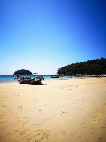 Longtail boats sit on the shoreline of the golden sands of Kata beach. The sea is a bright blue-green. 