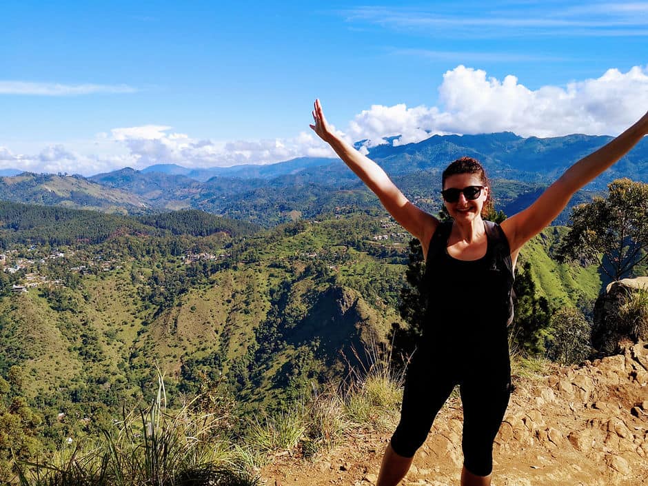 A woman stands on the top of Ella Rock mountain, surrounded by rolling hills covered in thick forest, in Ella, Sri Lanka