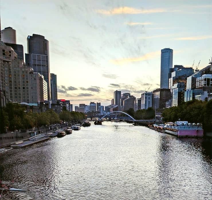 Standing over the Yarra River in Melbourne, either side sits high rise modern tower buildings with boats lining the water's edge. The sun sets in the distance