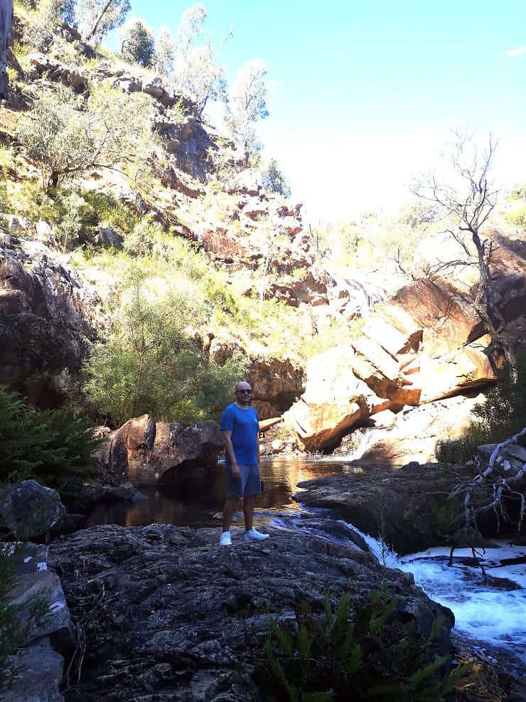 Standing on a larger rock sitting in the stream that flows from the McKenzie falls, a steep cliff sits behind the stream, shading the path