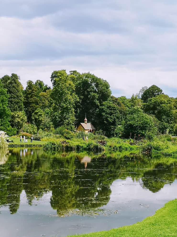 A small hut sits on the banks of the lake in Melbourne's Botanical Gardens, surrounded by large, leafy green trees, which reflect in the still water. 