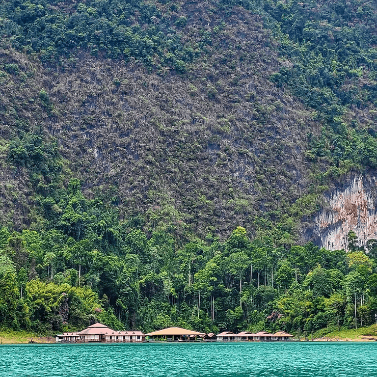 A huge wall of limestone cliffs sits behind a lake house, floating on Cheow Lan lake. The buildings look tiny in comparison to the scale of the cliffs