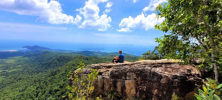 A man sits on a cliff edge at the top of Dragon Crest mountain in Krabi, Thailand, overlooking a thick green forest. 