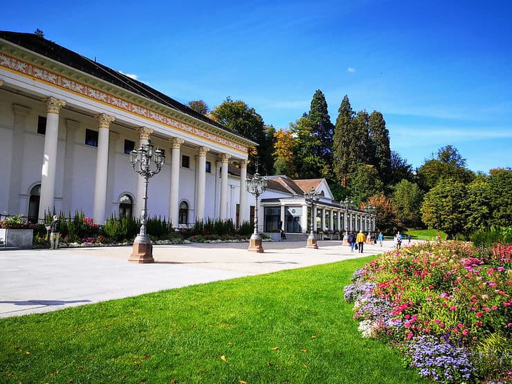 The grand casino in Baden-Baden. Gold leaf patterns surround the top of the building which has a series of pillars to the front. In front of the casino a neat lawn and a landscaped garden.