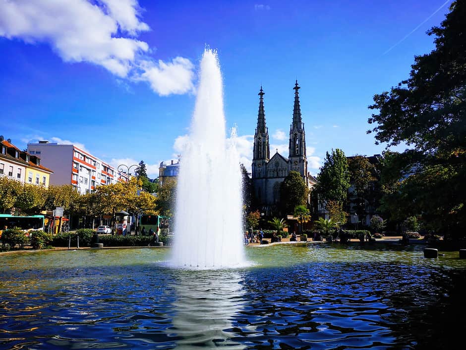 A water fountain sprays water high into the air from the centre of a man-made water feature. Surrounding the water are leafy trees and a church with two tall, gothic style turrets. Baden-Baden, Germany