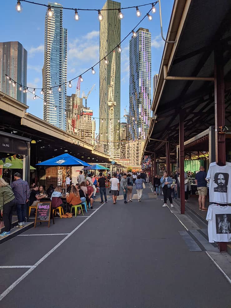 Covered market halls sit either side of a path in Melbourne's Queen Victoria Market. Strings of light bulbs hang of the roof of the markets, above the path and in the centre you can see tall skyscrapers from the city