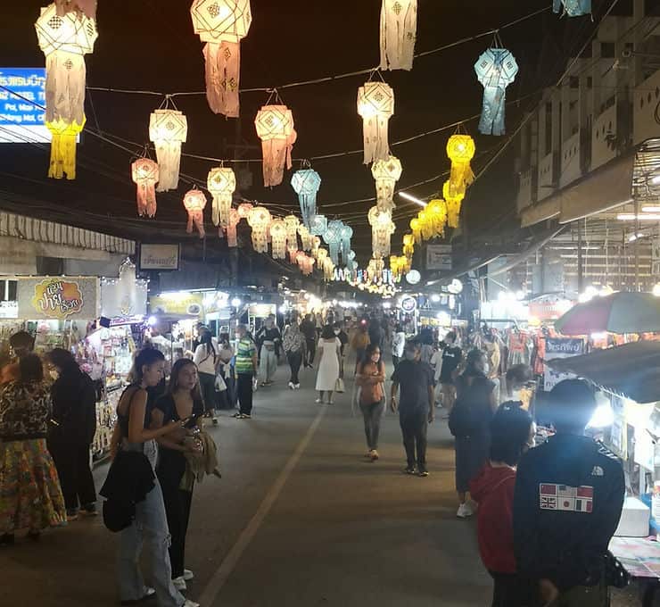 Strings of illuminated pastel-coloured lanterns hang over the centre of the street at night. Market stalls line either side of the street with lights shining from the stalls.