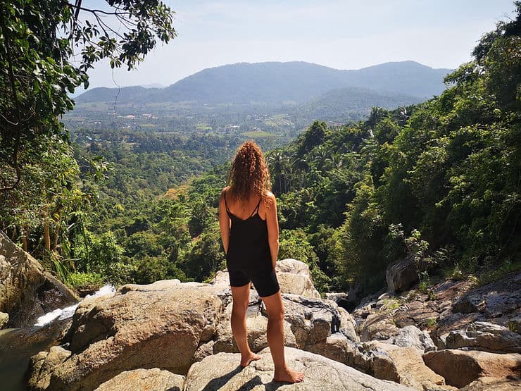 A woman stands barefoot on rocks at the top of Na Mueang waterfall in Koh Samui, Thailand, looking out to the landscape