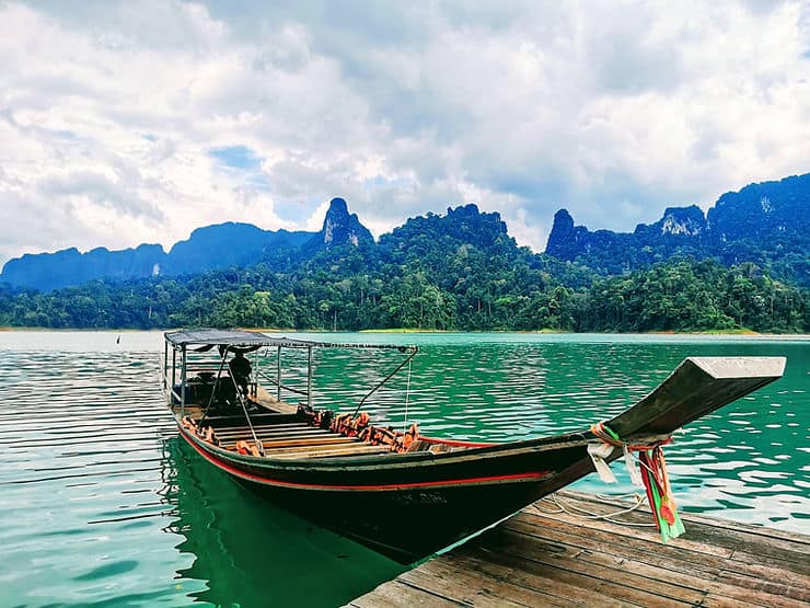 A long tail boat floats on the lake in Khao Sok National Park, Thailand, surrounded by thick jungle and limestone mountains