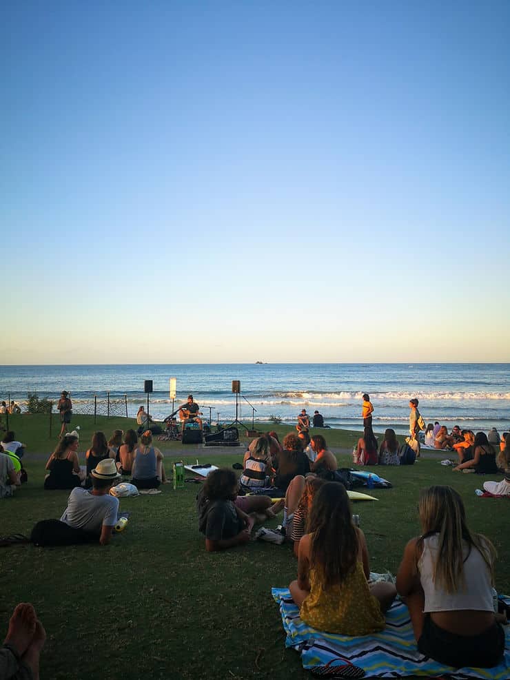 Small groups of people sit on the grass behind Main beach in Byron Bay, watching the sunset as a guitarist plays