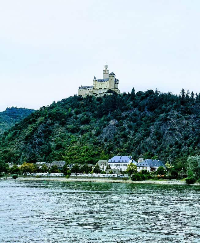 A stone castle rises high above the tree tops on a steep hillside, alongside the Rhine River in Germany.
