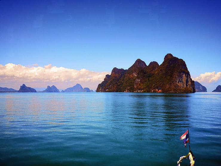 Jagged peaks of the Phang Nga Bay islands line the horizon. The sky and the sea are both deep blue and a small Thailand flag appears in the bottom corner of the image, from the front of the boat