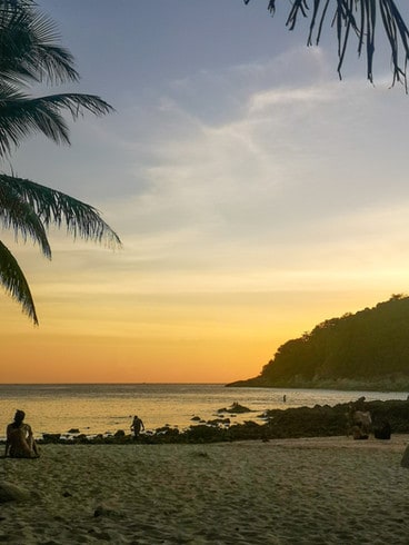 The sunsets over Yanui beach, and the sky turns bright orange Palm trees frame the photo and a small hill sits on one side. 