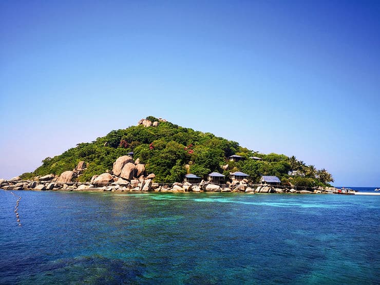 The small island of Nangyuan off the coast of Koh Tao in Thailand is covered in large boulders and green trees, surrounded by turquoise blue sea