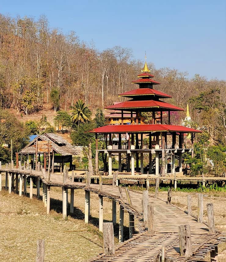 A bamboo bridge stands over a dry rice field, next to a red roofed pagoda and surrounded by a dry forest of trees, in Pai, Thailand