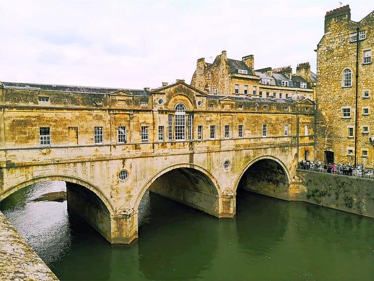 A thick stone bridge with three arches straddles the River Avon, in Bath, UK. The bridge houses a grand building with small glass windows.