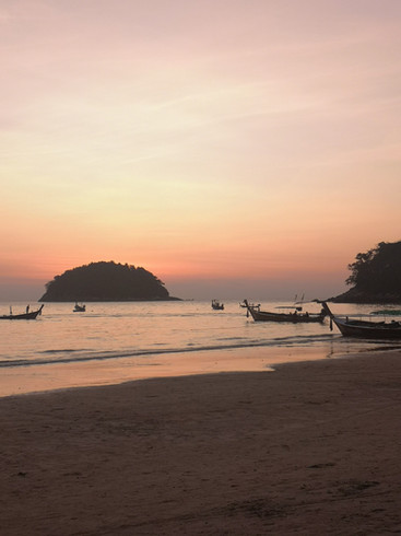 The sky glows orange-pink as the sun sets over Kata beach. Several longtail boats float in the shallow sea.
