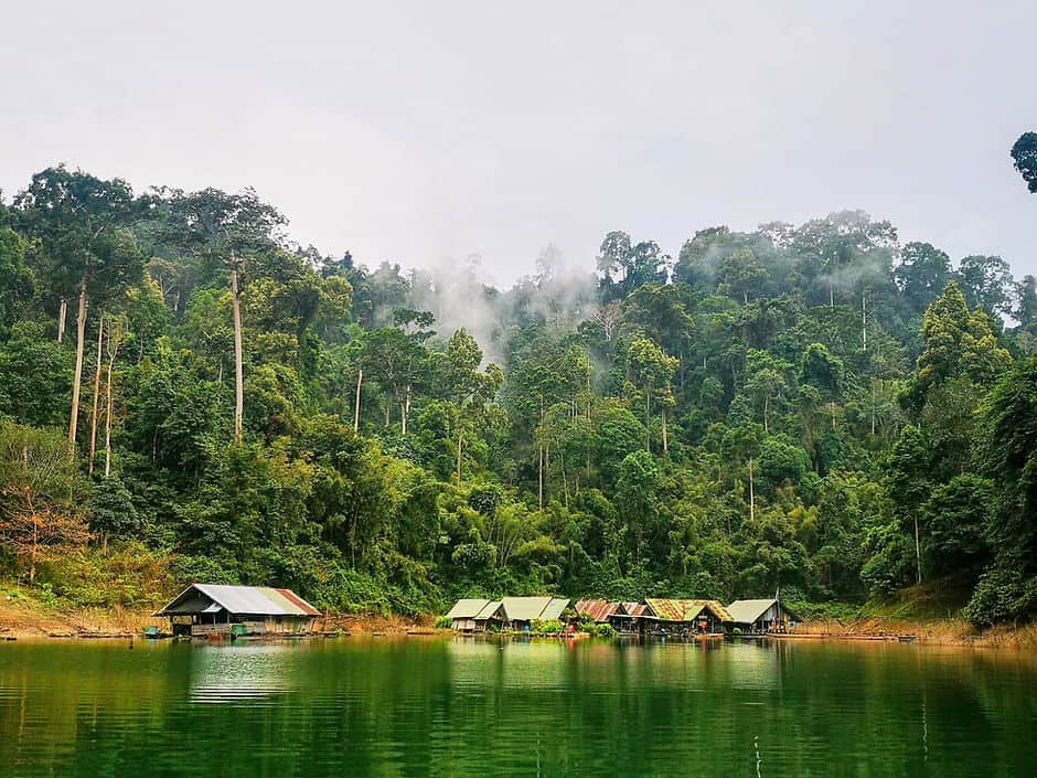 Small tin-roof shacks sit by the edge of a lake in Thailand. Steam rises from the thick rain forest behind.