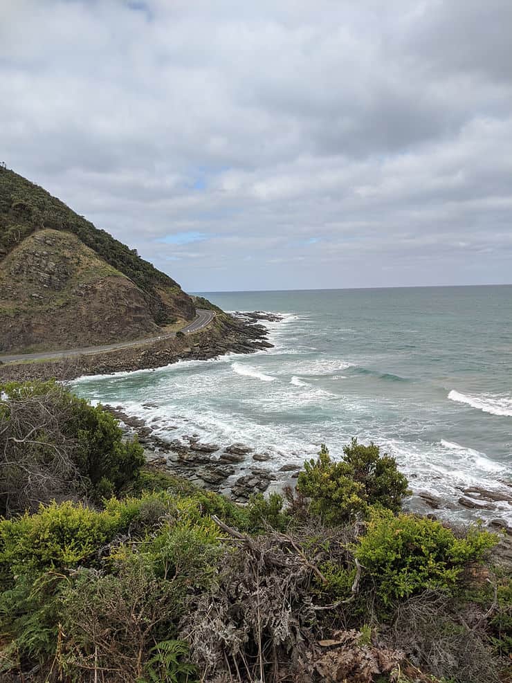 The Great Ocean Road winds gently around the curve of the mountain, nestled between the peaks and the ocean with waves crashing against the rocky shoreline