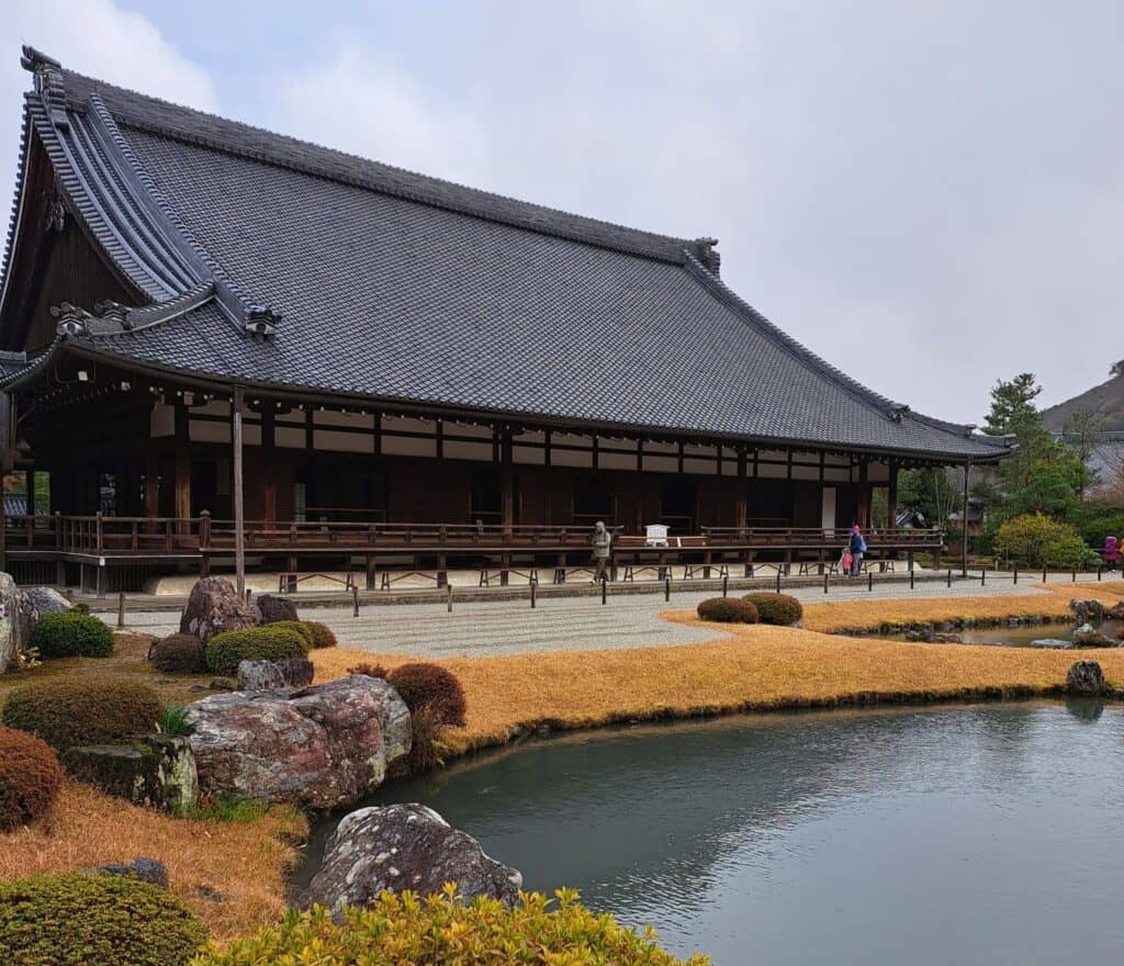 The large pitched roof of the Tenryu-Ji temple sits in front of a tranquil pond in Arashiyama, Japan