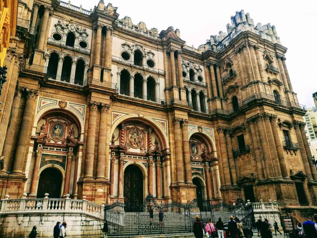 The historic cathedral of Malaga has large arched walkways and smaller arched windows on the second floor. A turreted roof runs all the way around.