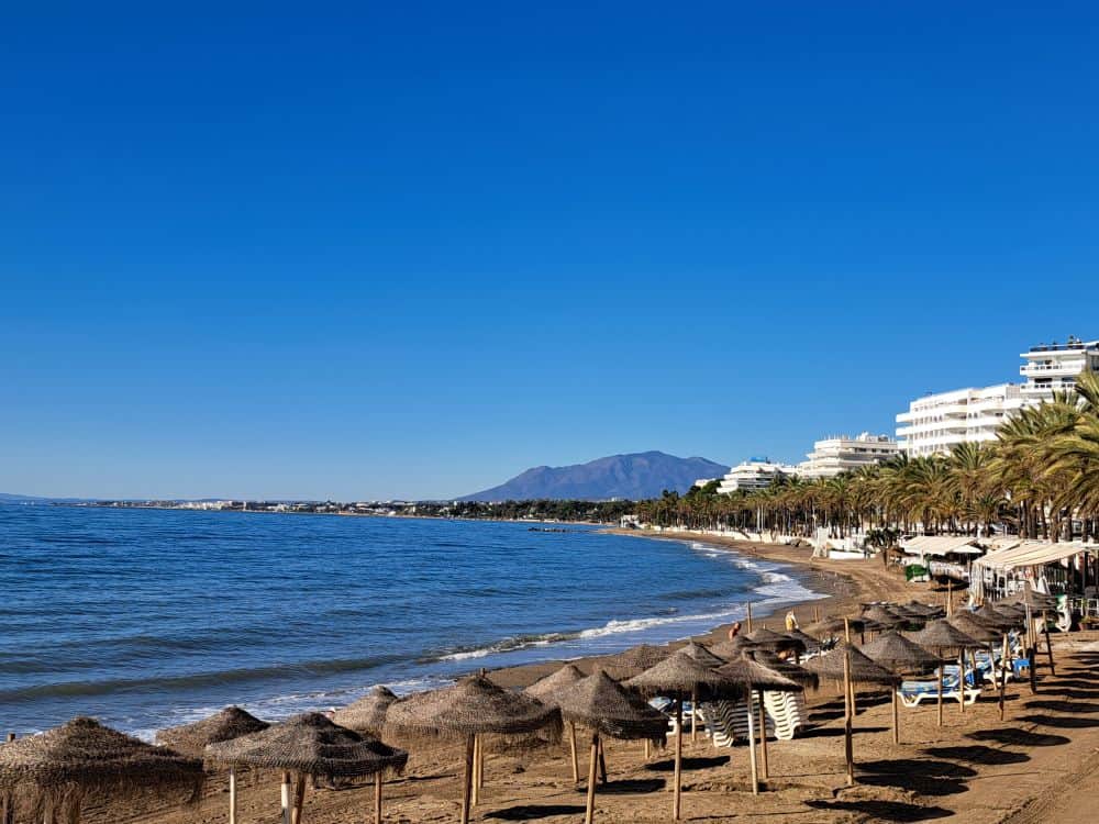 Golden sands of Marbella beach with straw umbrellas and sunbeds and mountains in the background.
