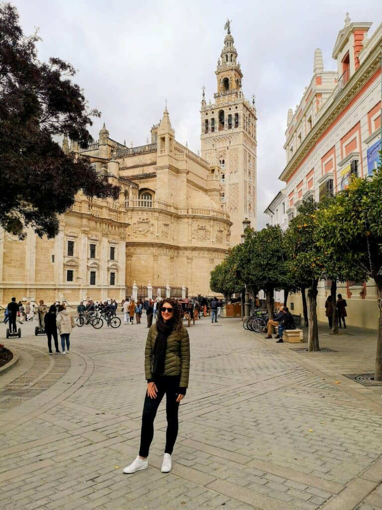 A woman stands in a square, in front of the impressive Seville Cathedral, with it's tall bell tower.