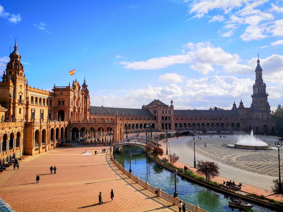 Seville's Plaza de Espana. A brick building is built in a semi circle, around a brick-paved promenade and thin waterway. Curved footbridge cross the water.