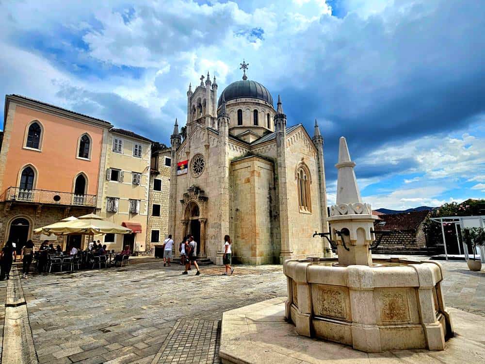 A historic water fountain sits in front of a small stone church in a quaint cobbled old town.