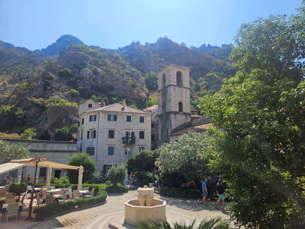 A stone clock tower and grand building sit in a square, with a stone water fountain, with tree covered mountains behind them.