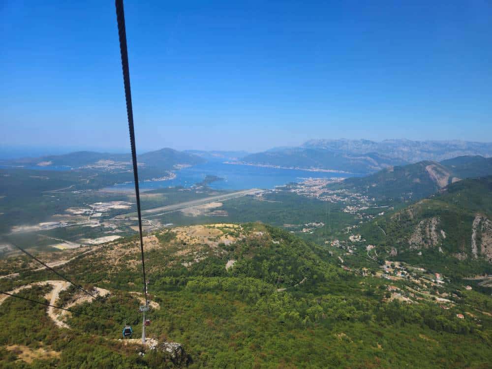 The images shows the view from inside a cable car. Below is green forest and a blue bay.
