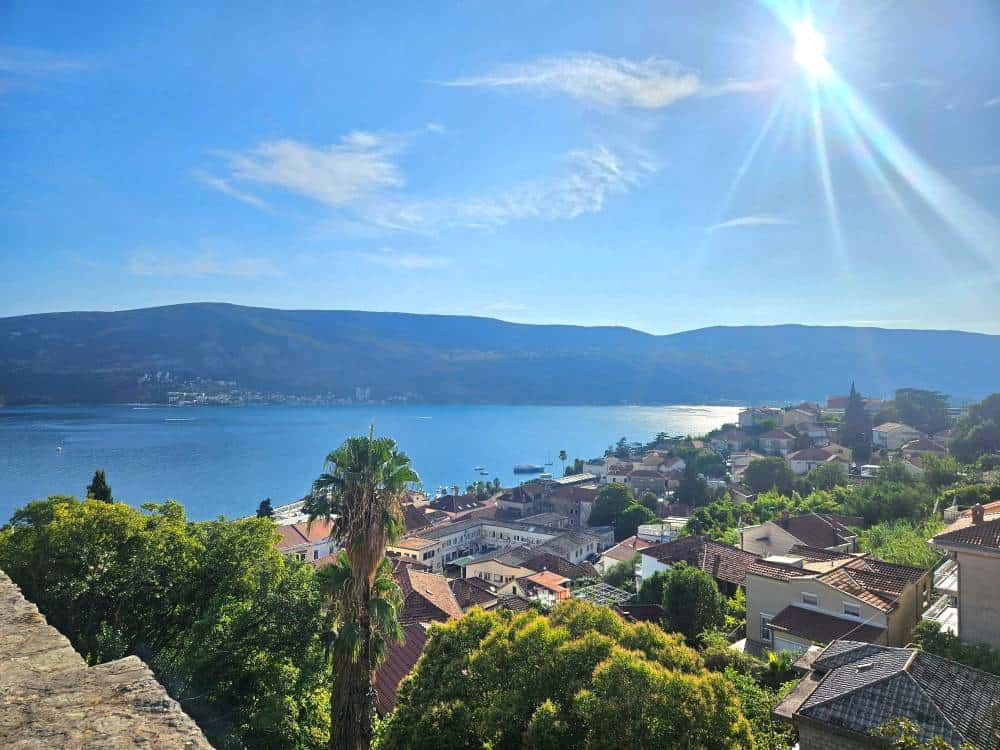 Looking down over terracotta rooftops and green palm trees, with a bright blue sea and distant mountain range.