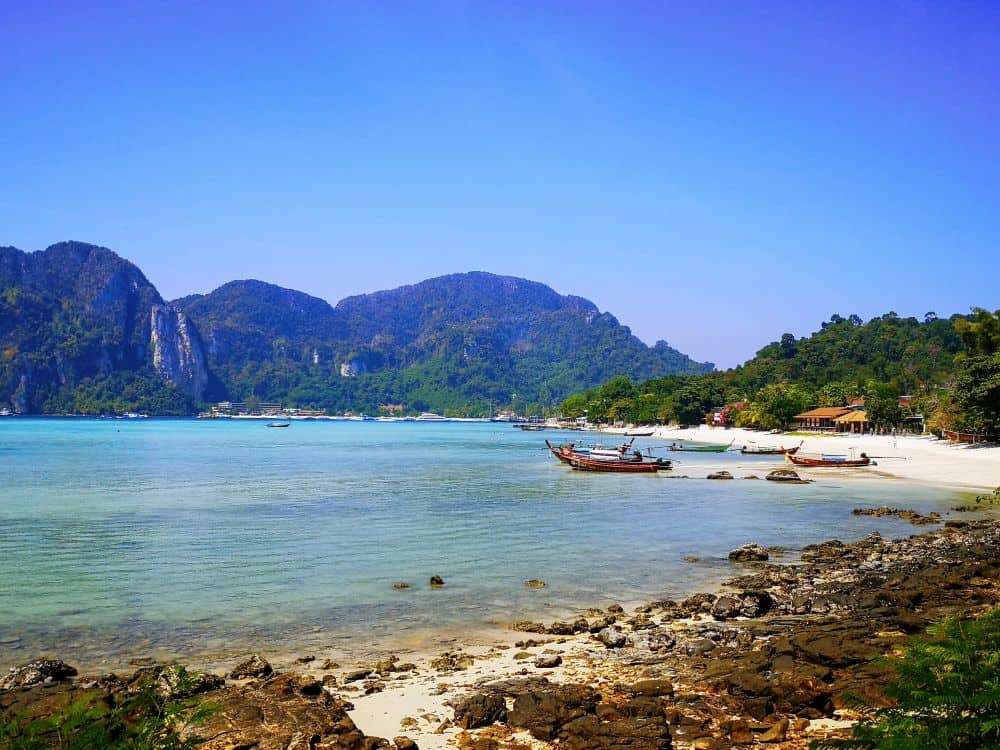 A single wooden longtail boat floats on a calm blue bay. In the background are tall limestone cliffs and at the shoreline of the bay are rocks and white sand.