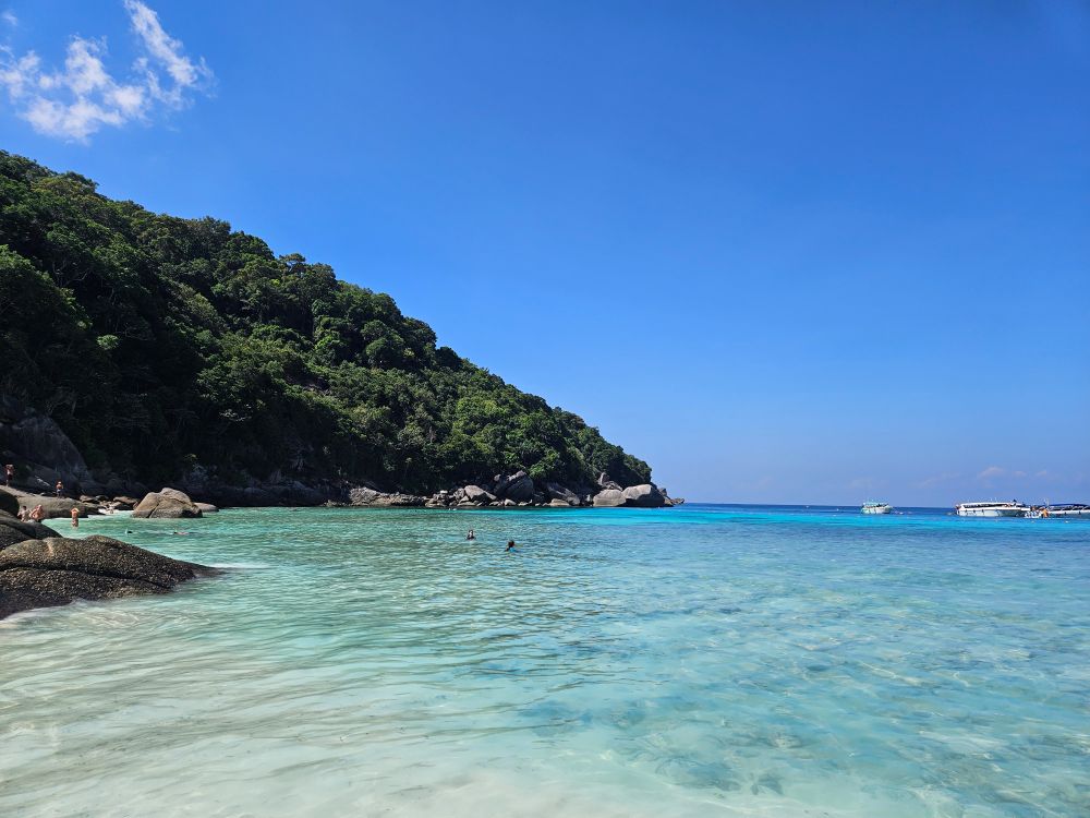 A shoreline with crystal clear sea water, and white sand. A tree-covered mound rises from the side of the bay.