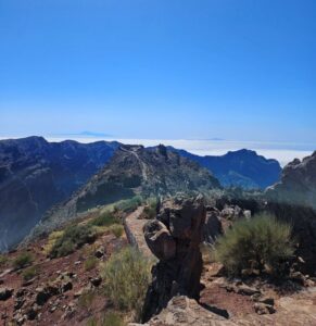 A path leads up to a viewpoint at the top of a mountain range, looking across mountains and the clouds below